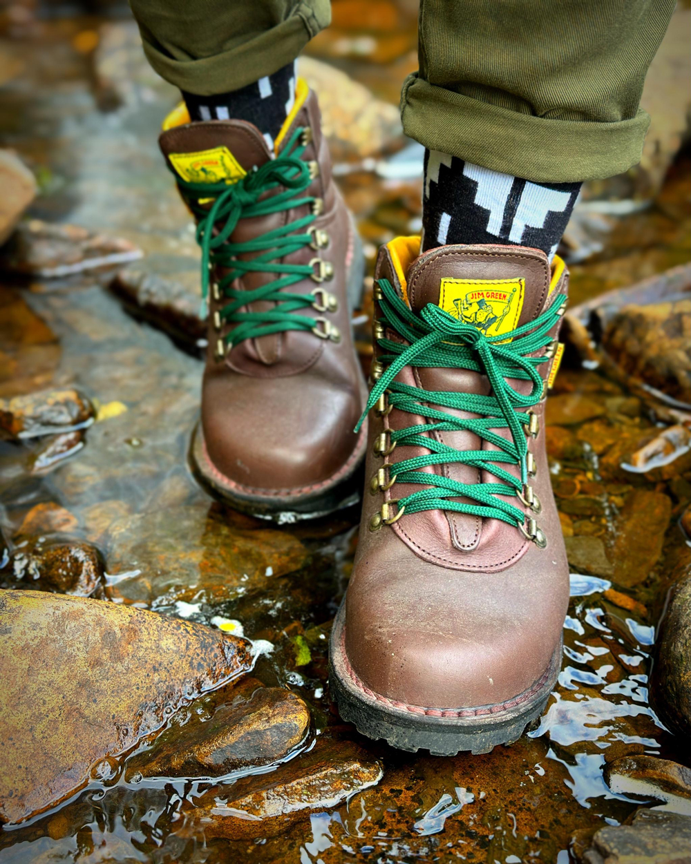 Close up of brown Jim Green boots with green laces standing in a shallow rocky stream